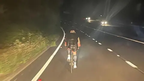 Amo Panesar Man in black cycling on a road at night-time. He is being followed by someone lighting his way from behind. There are cars on the opposite side of the road. A bank of grass is to the left side of the cyclist.