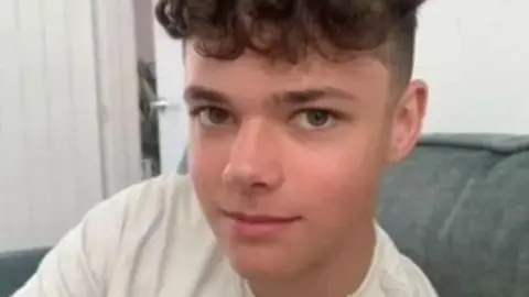 Family handout A teenage boy with brown curly hair, short on the sides. He is wearing a white Under Armour t-shirt and a thin chain as he sits on an armchair.