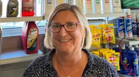 A woman with short blonde hair smiles while wearing purple framed glasses and a blue top with white details. Behind her is various stock from the petrol station including sweets, oil and de-icer 