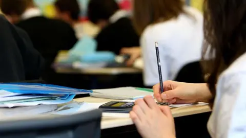 Getty Images Pupils in a classroom. In the foreground is the side of a pupil holding a pencil. There is a calculator, books and papers on the desk. 