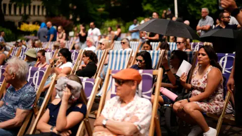 People sit in deckchairs during a hot, sunny day at the Victoria Embankment with some shielding themselves using umbrellas, paper fans, or clothing to cope with the heat.