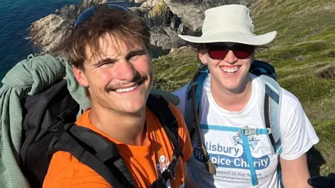 James Hirst Man and woman on a coastal path hiking. They are posing for a selfie and smiling, and have rucksacks on their backs