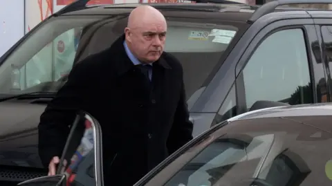 PA Bald man exiting a black car wearing a suit and black overcoat. 