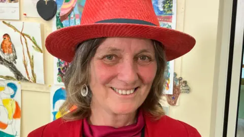 Joy Roodenburg-Vermaat. She is wearing a red straw hat which has hair sewn in. Joy is looking directly at the camera and smiling.