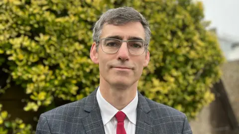 Deputy Montfort Tadier smiling at the camera. He is wearing a checkered blazer, a white shirt and red tie. He also is wearing glasses and is stood in front of a green bush.