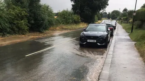 RICHARD HARRISON This is a picture of the main road near the housing site, where campaigners say the drains are already struggling to cope. It shows five cars parked at the side of the road, with surface water flowing underneath them and across part of the road. A 30mph sign is attached to a lamp-post on the right of the image, a thick hedgerow is on the left.