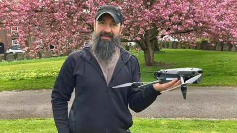 A man with a hat and long beard stands and holds a drone in one of his hands. There is a large tree behind him.