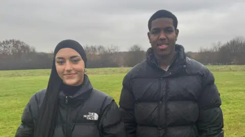 Zainab usa um hijab preto e uma jaqueta North Face preta. Jaiden está vestindo um casaco preto. Eles ficam um ao lado do outro em um campo com árvores ao longe. 
