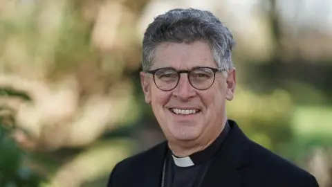 Diocese of Worcester A man with short, slightly wavy greying hair is wearing round, dark-rimmed glasses, a clerical collar with black shirt and jacket, and standing in front of a blurred background of trees outside.