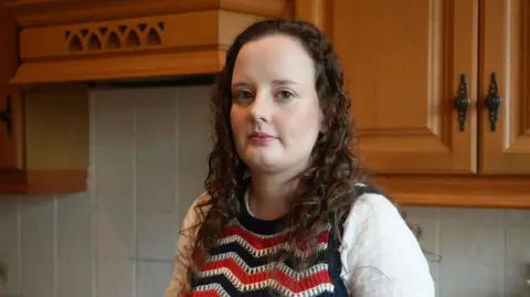 BBC Katherine Jauncey is looking at the camera. She is stood in her kitchen, in front of wooden cabinets. She is wearing a white shirt and red, white and navy knitted vest.