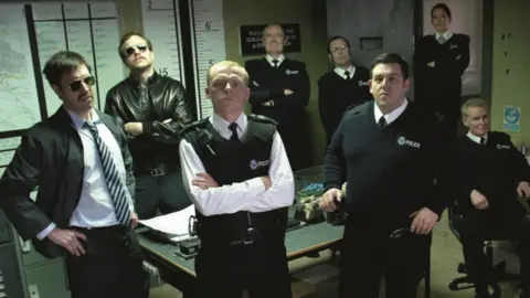 The actors from Hot Fuzz posing in police uniforms inside a police station. They are looking into the camera.