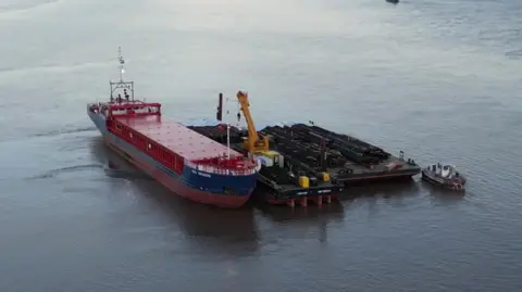 A red and blue cargo ship floats alongside tug boats, with a crane removing cargo