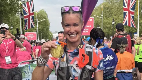 Emma Holmes A woman holding a gold medal on the finish line, smiling after completing a race. You can see other runners in the background. 