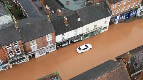 Aerial shot showing brown floodwater along a main shopping street with an abandoned white car in the middle 