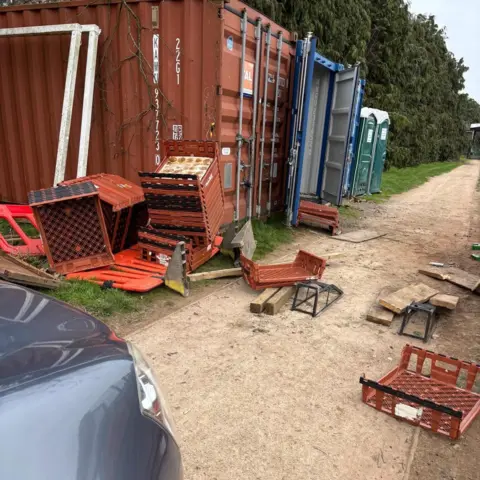 Mostyn Rangers FC A brown and a blue shipping-style container on a patch of grass next to a dirt path. Boxes and broken crates are strewn across the path.