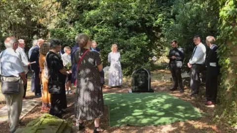 A group of people are gathered around a grave in sun dappled light. In the centre is a black marble headstone with light grey engraving