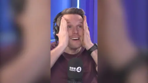 Greg Foot clasps his head in his hands and looks worried. He is wearing a brown T-shirt and is sat behind a Radio 4 mic.
