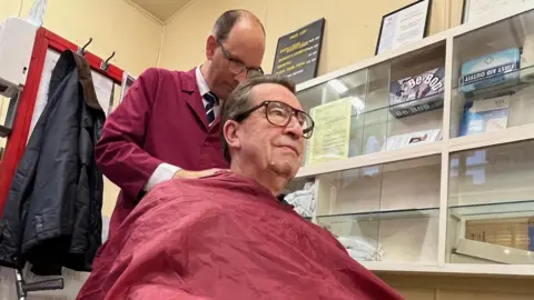 Kevin Shoesmith/BBC Customer David Jackson, covered with a maroon cloak, is pictured having  the back of his hair cut by Mr Hutchinson. A crutch belonging to Mr Jackson is seen on the left of the image.