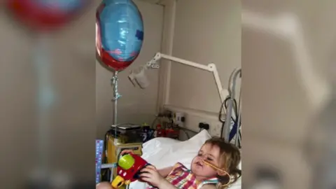 Walls Family Emily as a young child in a hospital bed with a drip connected to her nose. She is playing with a toy and has short light hair. 