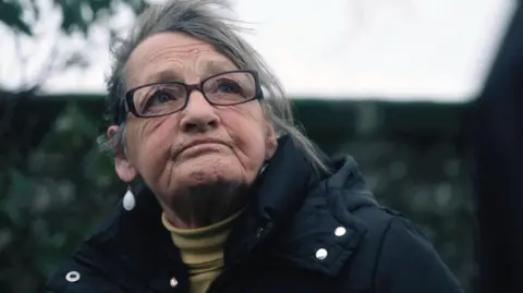 A woman wearing glasses and a dark coat outdoors, seen from a low angle with trees in the background.