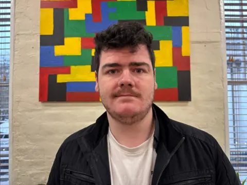 Man in a black coat and white top looks at camera. He has dark hair and stubble. A piece of brightly coloured artwork hangs on the wall behind him. 