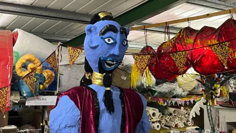 A large blue statue of the genie from Aladdin seeming made out of papier mache standing in a warehouse surrounded by other items including Chinese lanterns, rolls of fabric and gold and black bunting.