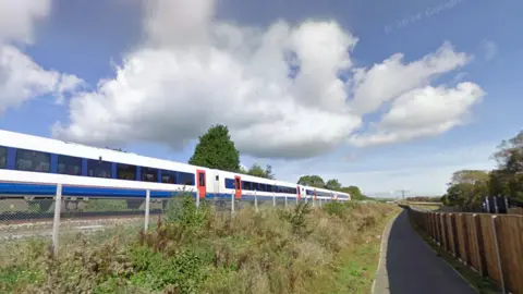 Google A blue and white train with red doors passes a section of public footpath