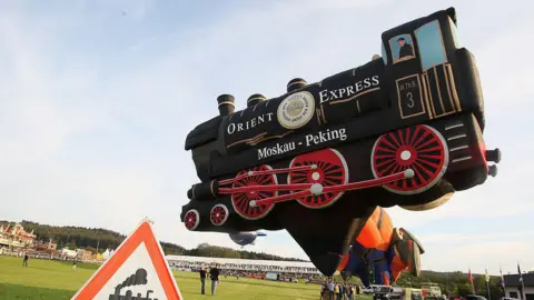 Getty A black train with red wheels hot air balloon that is tethered near a green field. 