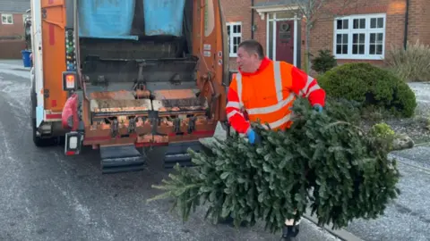 BBC A man in a bright hi-viz jacket is lifting a large Christmas tree into the back of a wagon. The vehicle is parked outside houses 