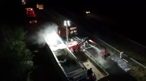 A night-time clean-up operation on a dual carriageway, viewed from an elevated position. A piece of heavy machinery with a lifting arm is positioned alongside the trailer and appears to be removing or handling material from inside it.