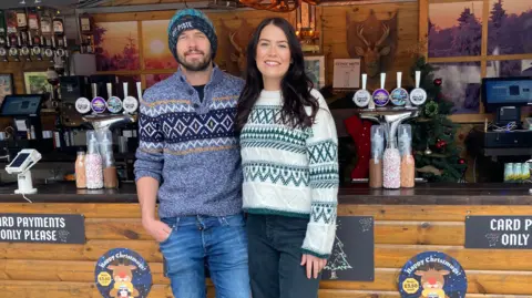 Abbey Matthews and her partner Kyle standing in front of one of their Bar At Yours stalls