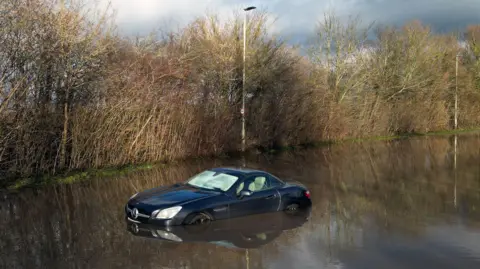 Joe Giddens, PA A black car is stranded in floodwater surrounded by trees during a storm in the UK in January 2025