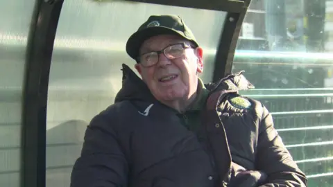 Peter Kirkley in a green baseball cap and wearing a thick black coat talks to the camera. He is also wearing glasses.