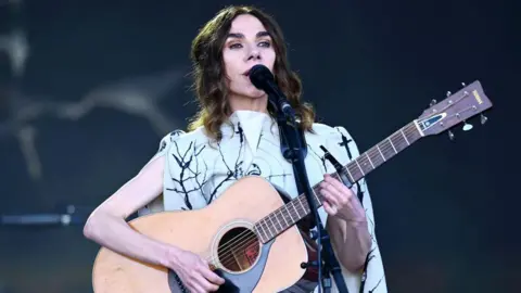 Getty Images A woman with brown hair and wearing a black and white dress playing a guitar and singing into a microphone.