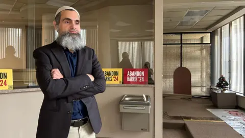 Hassan Abdel Salam stands with a blue sports jacket and short, and tan pants with his arms crossed. There are "Abandon Harris '24" posters behind him as well as a water fountain.