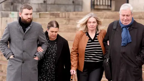 Joe Giddens/PA Wire A group leaving the court. From left to right: A tall man in his 30s with a brown beard, wearing a long straight grey coat. A woman of a similar age, up to his shoulders, wearing a black dress with some white patterns and a long black coat. A blonde woman in her 50s, wearing a black and white striped top and brown leather jacker. A man who looks in his 60s with white hair, wearing a long black coat and blue scarf with a black satchel over his shoulders. Each couple has their arms linked.