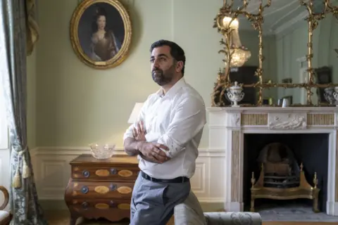 PA Media Yousaf standing in an elegant, traditionally decorated room with arms crossed, positioned near a fireplace with ornate gold-framed mirrors and a wooden chest of drawers beneath a portrait on the wall.