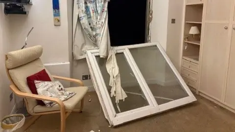 The UPVC window that was blown in to a room during the storm. It is whole but has ripped the curtains and it is resting on the carpet. There is a chair, a TV and a wardrobe unit in the room.