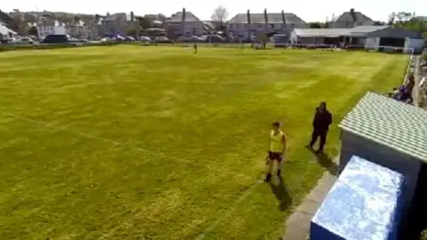 A football pitch with two men on the sideline, one is wearing a black coat and the other is a linesman in hi-vis