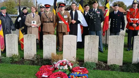 Crown Copyright Seven identically shaped headstones in a row, facing forwards, engraved with a cross and words. Wreaths of poppies are laid in front of the newest grave, and behind the graves stand a group of people. One is a vicar wearing white robes and a black cassock, others are wearing brown or black miltary uniform and holding flags. One man appears to be a family member, standing holding an order of service.
