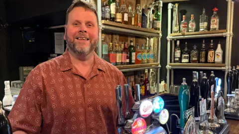 A man stands behind a bar wearing a patterned shirt