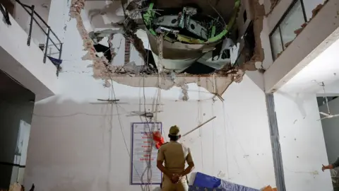 An official looks up with his arms behind his back and back turned to the camera, as they inspect a huge hole in the wall of a building interior, with some of the plane visible in the hole, on Thursday.