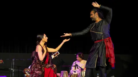 Vipul Sangoi Two dancers on the stage, both are performing Kathak while wearing traditional Indian clothes. A tabla (drums) player is in the background 