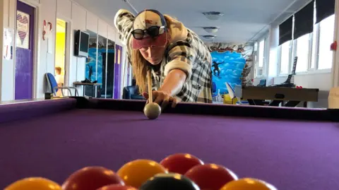Jon Wright/BBC A young man with long hair and a cap lines up a break on a pool table