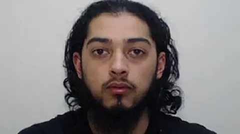 Greater Manchester Police A mugshot of a young man with long dark hair and a dark beard. He has a blank expression on his face.