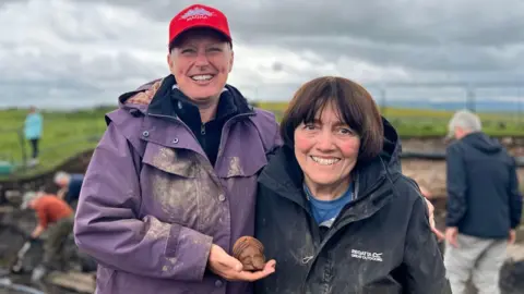 The Vindolanda Trust Rinske de Kok and Hilda Gribbin are both smiling widely and dressed in dusty coats at the archaeological dig site. They are holding the terracotta head in front of them.