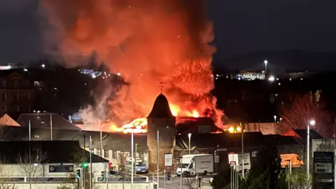 Iona Paton Night time shot of fire ripping through a warehouse building roof in an industrial estate. Flames and smoke are rising from the roof of the building which has a brick tower and domed roof at one corner.