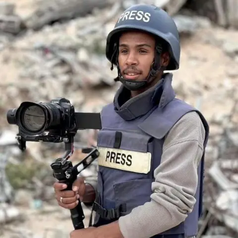 Reuters A man wears a press vest holding a camera and wears a helmet. He is stood infront of rubble