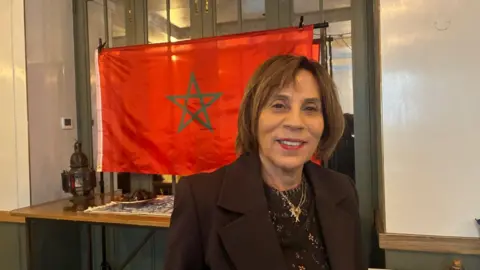 Liliane Harris is seen with short brown hair wearing a dark blazer, smiling in front of a red Moroccan flag with a green star.
