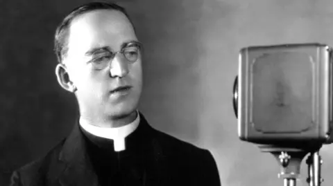 In an old black and white photograph, a priest , dressed in clerical garb, stands to the left of an old style radio microphone 